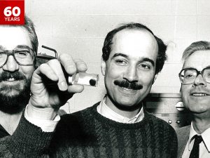 Black-and-white photo of Bozidar Mitrovic, Fereidoon Razavi and the late Frans Koffyberg in which Razavi (centre) is holding up an object containing a circular blob; the other two men are looking at the object and smiling.