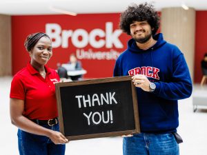 Two university students hold a chalkboard between them with the words "thank you" written on it in chalk.