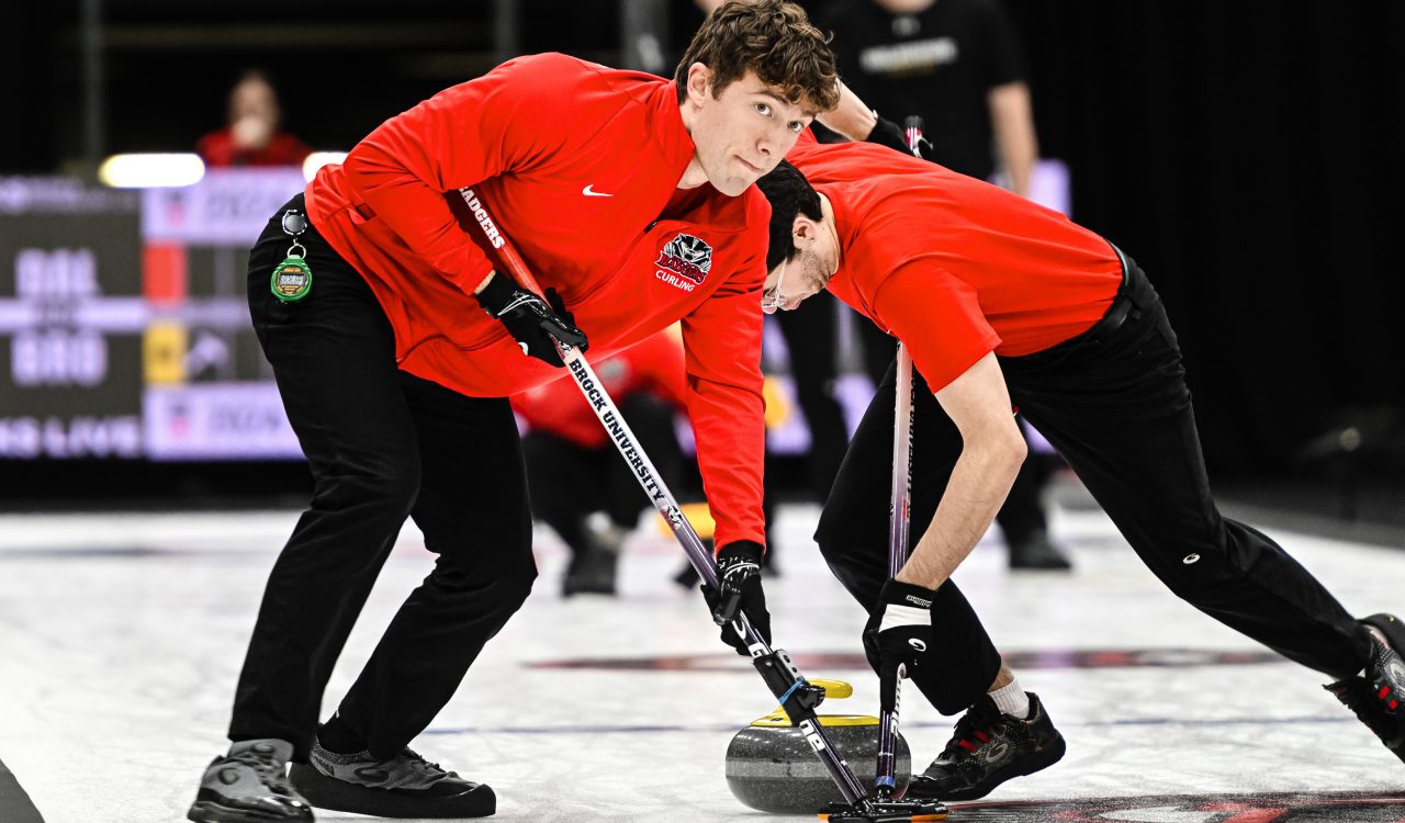 A man in red sweeps a sheet of ice with a broom during a curling match with a teammate sweeping close behind.