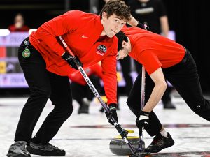 A man in red sweeps a sheet of ice with a broom during a curling match with a teammate sweeping close behind.