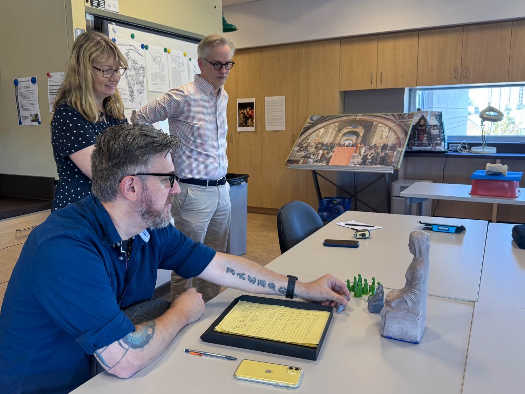 Three people stand in an archaeology lab looking at small figures.