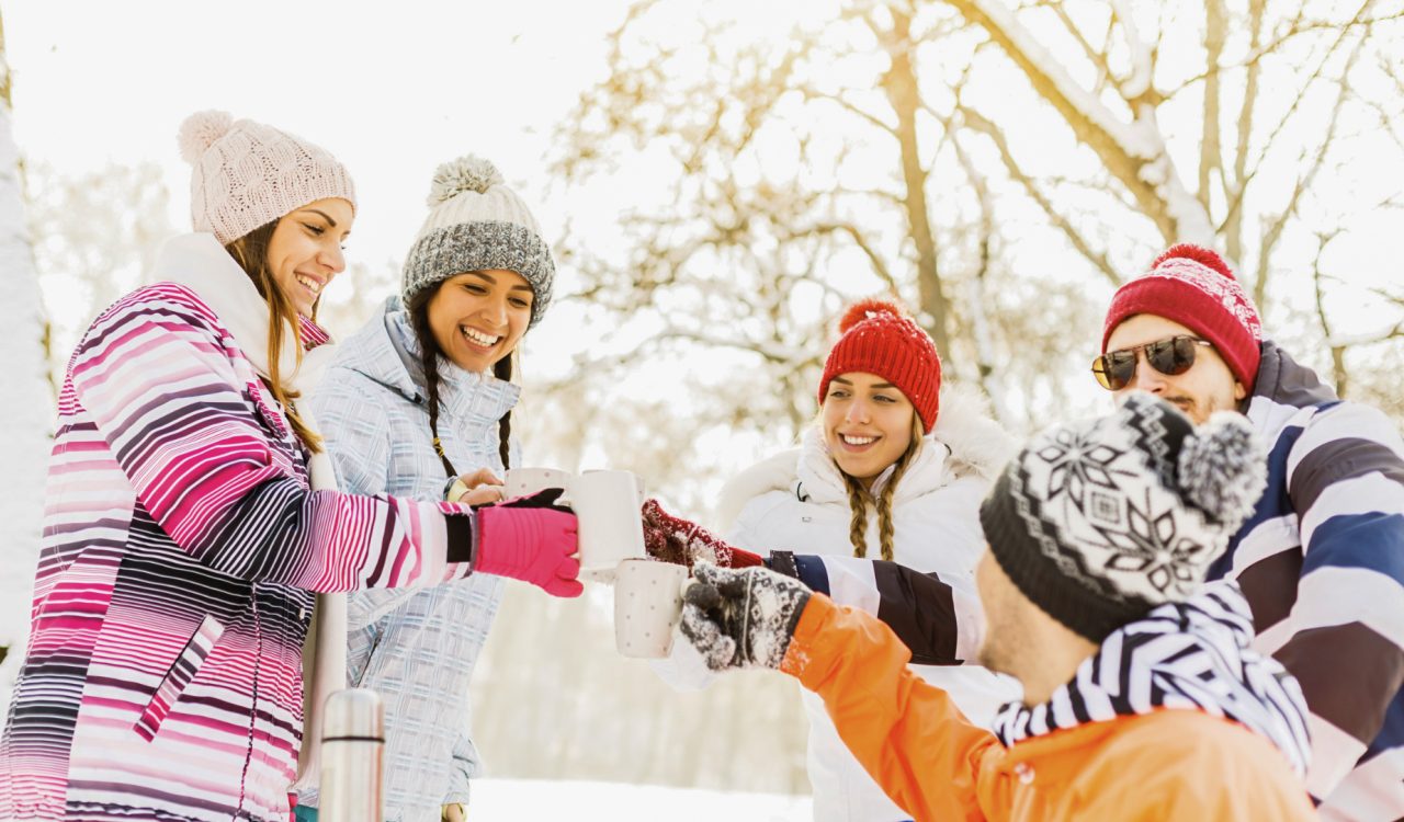 A group of friends in winter clothing toasting with hot drinks outdoors in the snow on a bright winter day.