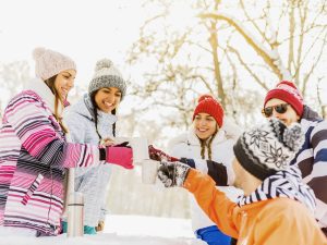 A group of friends in winter clothing toasting with hot drinks outdoors in the snow on a bright winter day.