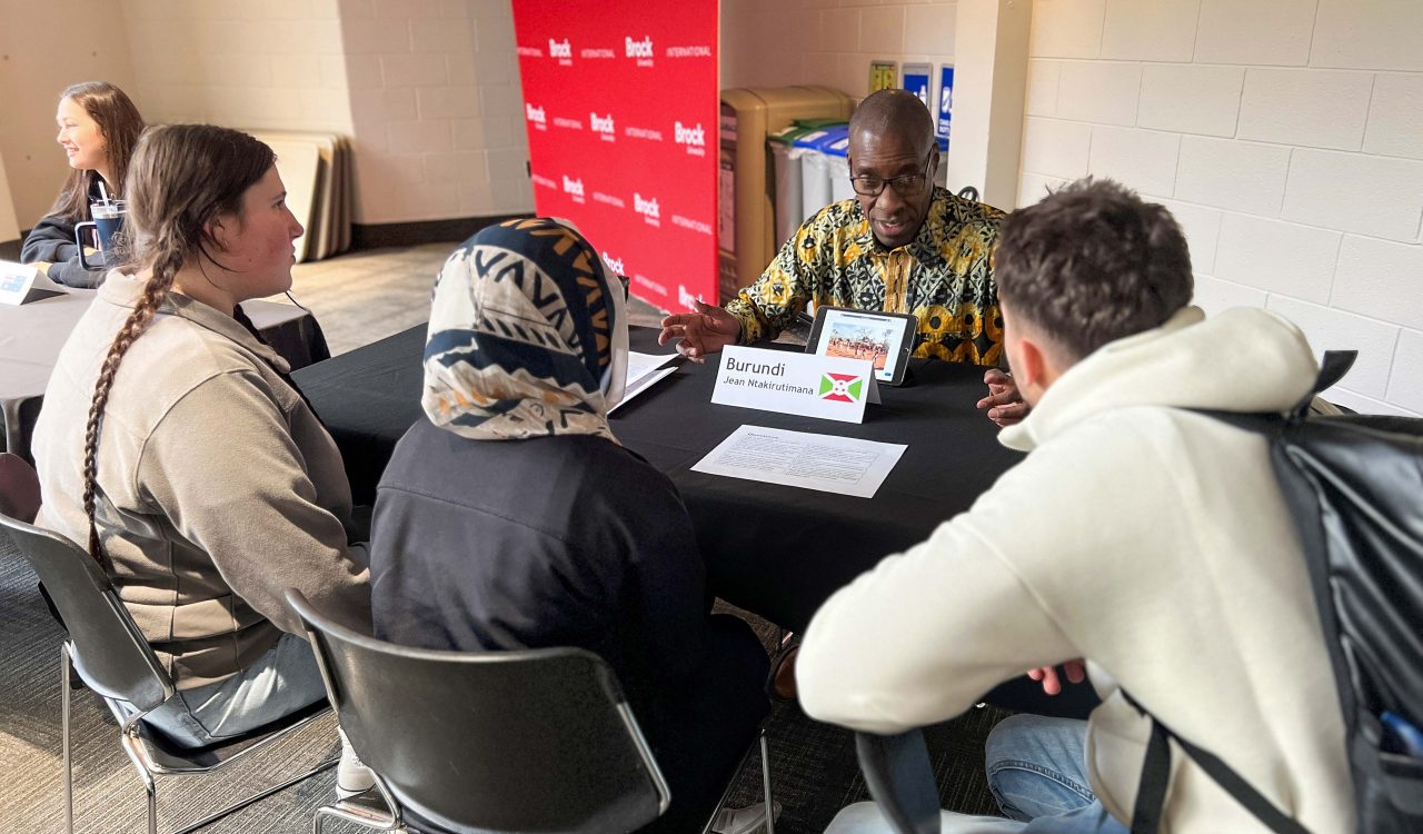 Three students sit around a table listening to a man from Burundi wearing a patterned shirt, engaging in a discussion during a cultural exchange event at Brock University.