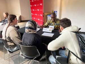 Three students sit around a table listening to a man from Burundi wearing a patterned shirt, engaging in a discussion during a cultural exchange event at Brock University.