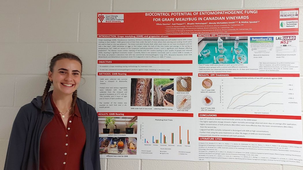 A smiling Olivia Devries stands next to a red-and-white poster with photos and words describing the research, hanging against a plain white brick wall.