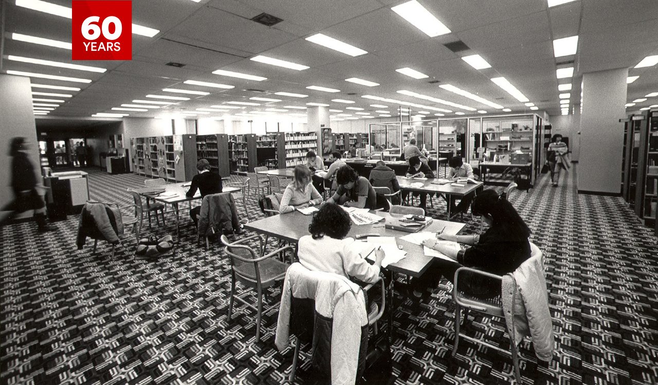 A black and white photo of students seated at study tables in the Brock University library during the 1980s while surrounded by patterned carpet and large bookshelves.