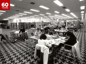 A black and white photo of students seated at study tables in the Brock University library during the 1980s while surrounded by patterned carpet and large bookshelves.