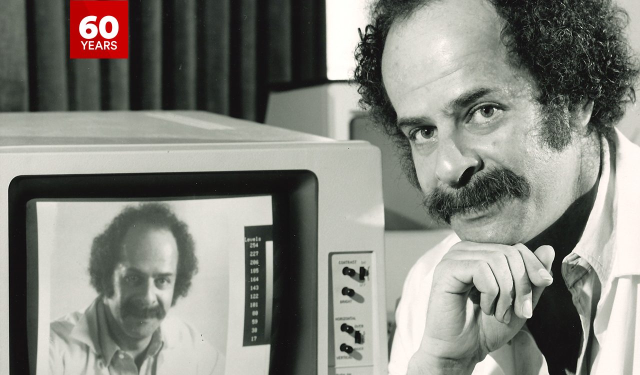 Close-up of then-Professor of Psychology Peter Ramm looking into the camera, head resting on his hand, with a machine and screen behind him; the screen is displaying his face.