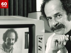 Close-up of then-Professor of Psychology Peter Ramm looking into the camera, head resting on his hand, with a machine and screen behind him; the screen is displaying his face.