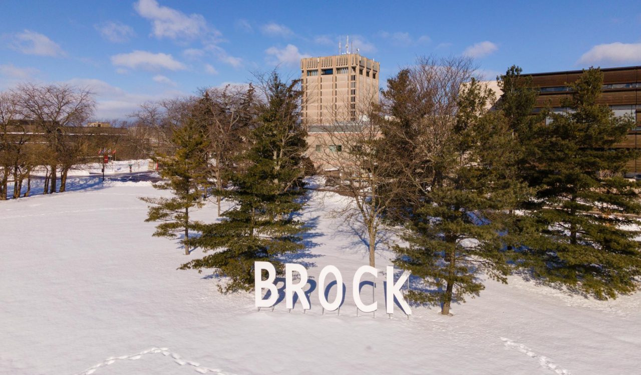 Giant letters spelling "Brock" stand in a snowy field in front of trees and a concrete tower.