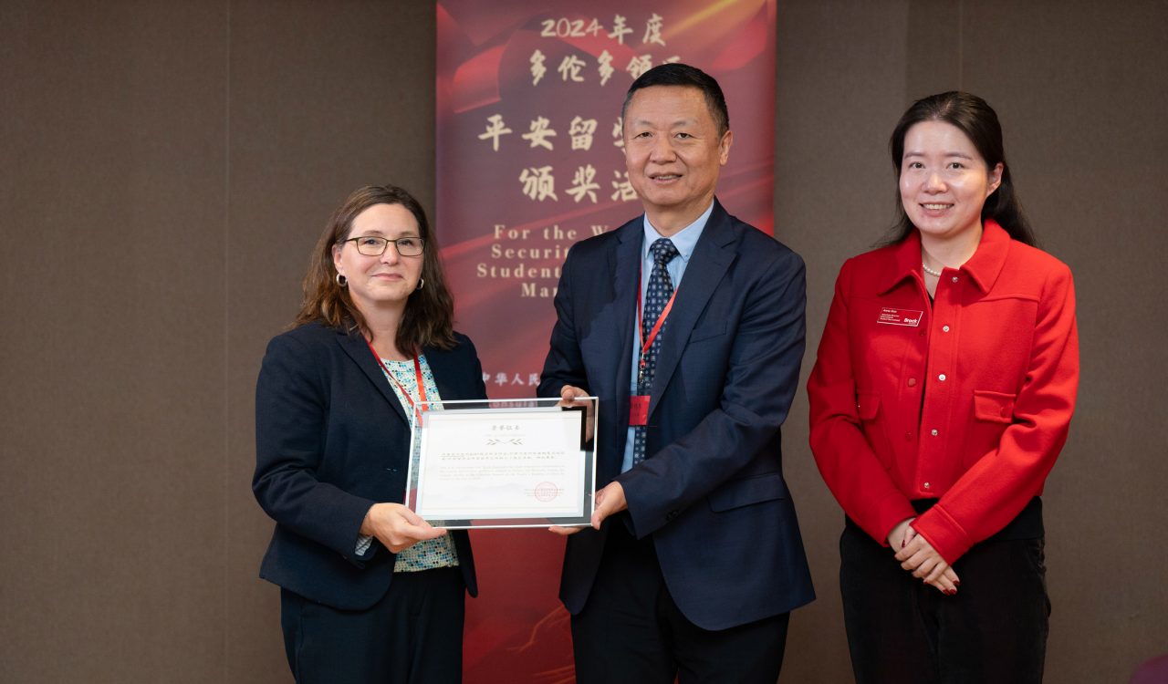 Three people in business casual attire pose for a photo in front of a Chinese banner. The man in the centre hands a woman to his right a glass-framed certificate.