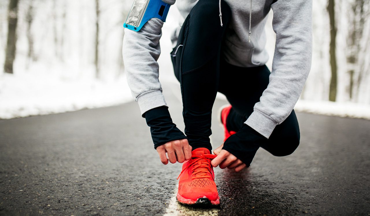 Person wearing running apparel kneels on pavement while lacing up an orange running shoe with snow on the ground in the background.