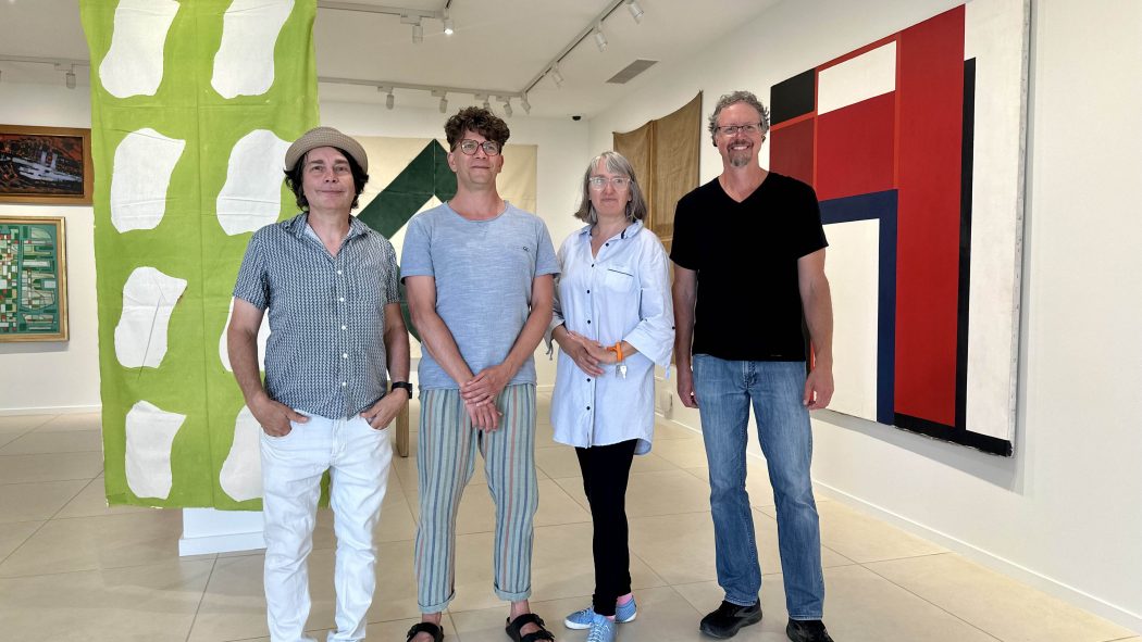 A group of four people stand shoulder to shoulder smiling at the camera in a brightly art gallery with paintings on the walls.