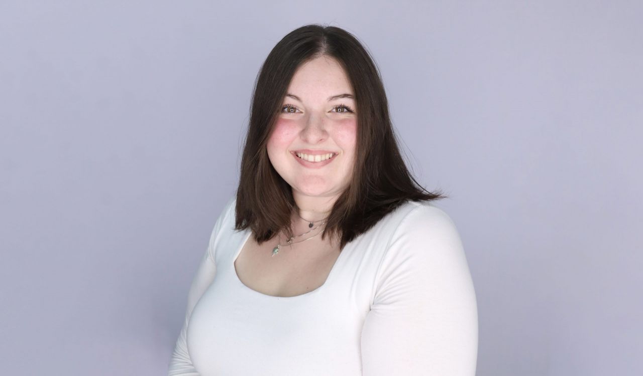 Ashley Giroux wears a white shirt and necklace and stands in front of a neutral-coloured wall.