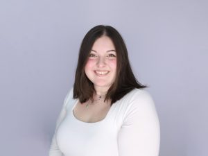 Ashley Giroux wears a white shirt and necklace and stands in front of a neutral-coloured wall.