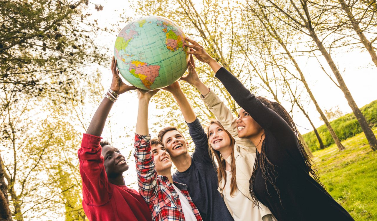 Young people stand in a circle holding up a globe in front of group of trees and blue sky.