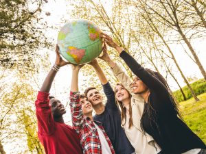 Young people stand in a circle holding up a globe in front of group of trees and blue sky.