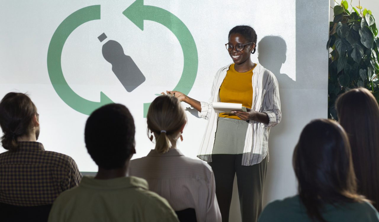 Young woman presents to a group of professionals with green recycling symbols projected on the screen behind her.