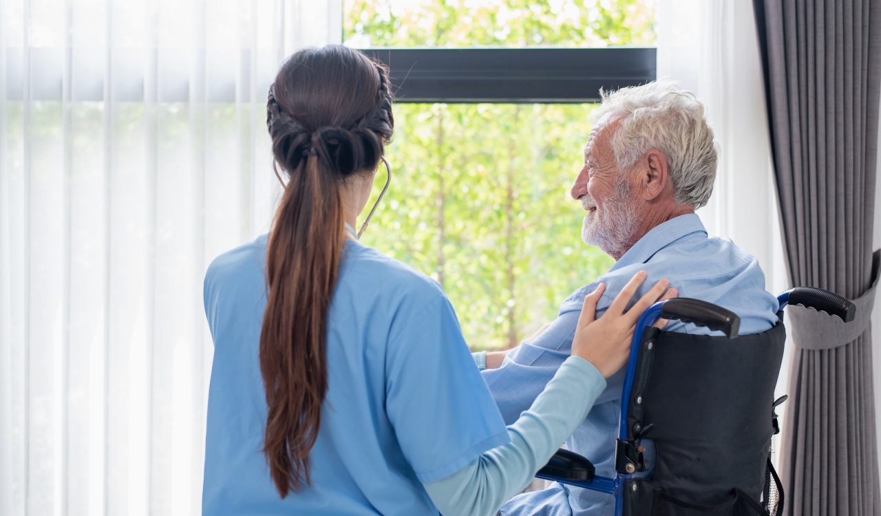 An older man, sitting in a wheelchair, looks out the window while a woman with long hair in a healthcare uniform, her back to the camera, stands beside him, her arm on his shoulder.