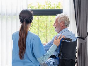 An older man, sitting in a wheelchair, looks out the window while a woman with long hair in a healthcare uniform, her back to the camera, stands beside him, her arm on his shoulder.