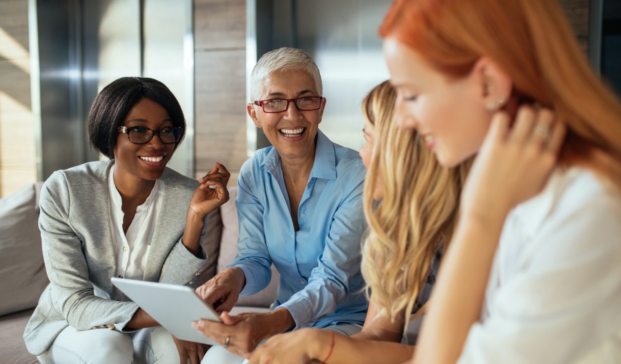 Four professional women smile and chat on a couch in front of a bank of elevators in an office.