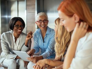 Four professional women smile and chat on a couch in front of a bank of elevators in an office.