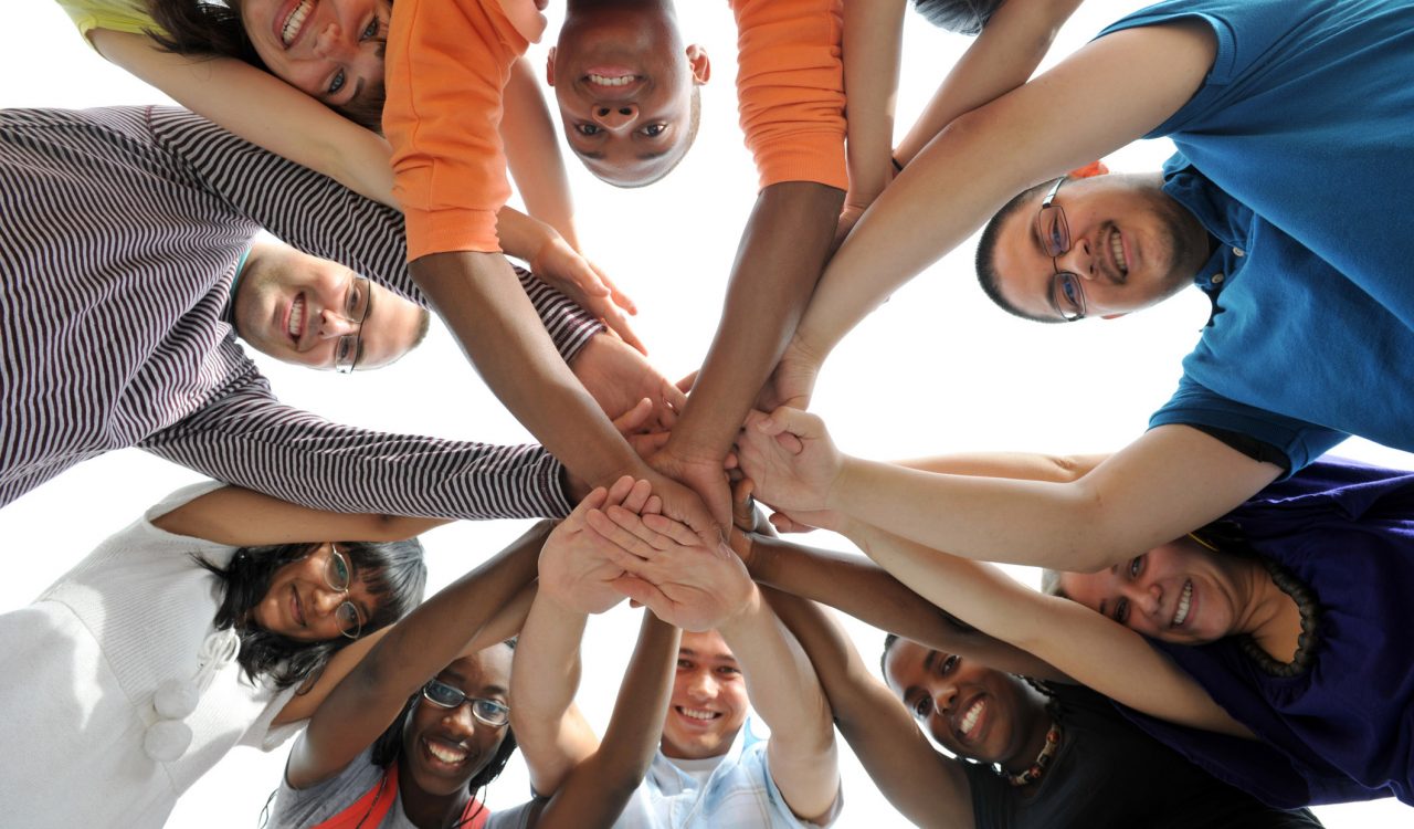 A worm’s-eye view of a group of teens putting their hands together in the middle of a circle.