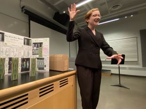 A young woman stands in a classroom in front of a large poster board presentation.