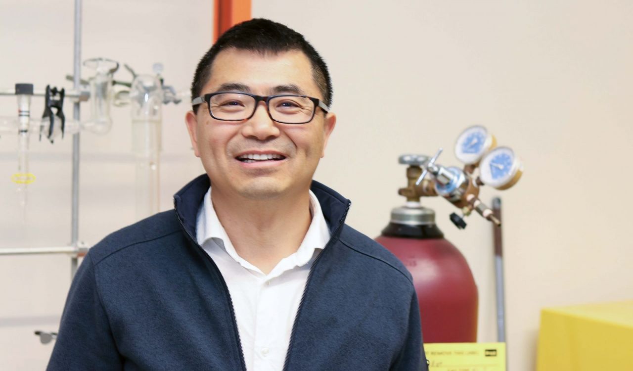 Close-up of Associate Professor of Chemistry Jianbo Gao smiling into the camera, standing in front of a tank and laboratory equipment hanging from a stand.