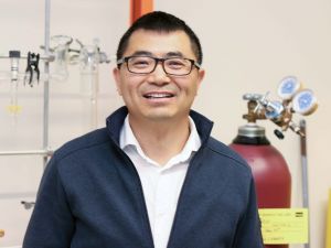 Close-up of Associate Professor of Chemistry Jianbo Gao smiling into the camera, standing in front of a tank and laboratory equipment hanging from a stand.