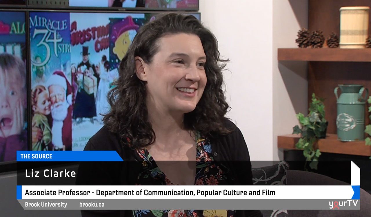 Woman with dark curly hair does a television interview sitting in a chair with christmas movie posters in the background.