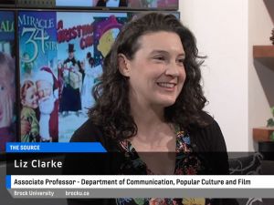 Woman with dark curly hair does a television interview sitting in a chair with christmas movie posters in the background.