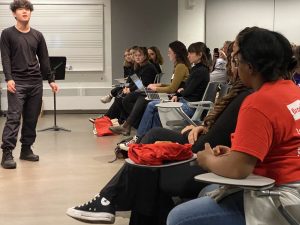 A young man stands in a classroom performing for a group of students sitting in chairs.