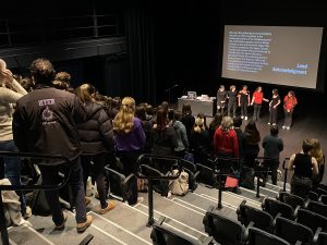 A large group of people stand in a theatre with banked seating looking at a smaller group of people standing on the black stage.