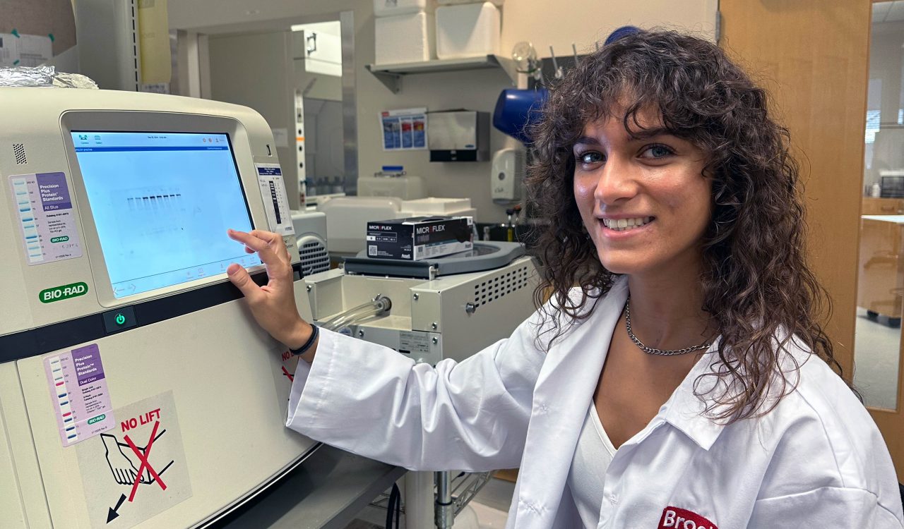 A young woman in a white lab coat smiles while pressing the screen on piece of lab equipment.