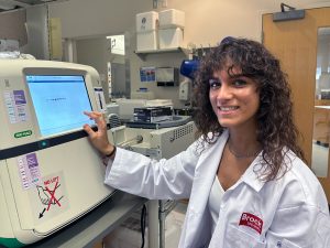 A young woman in a white lab coat smiles while pressing the screen on piece of lab equipment.