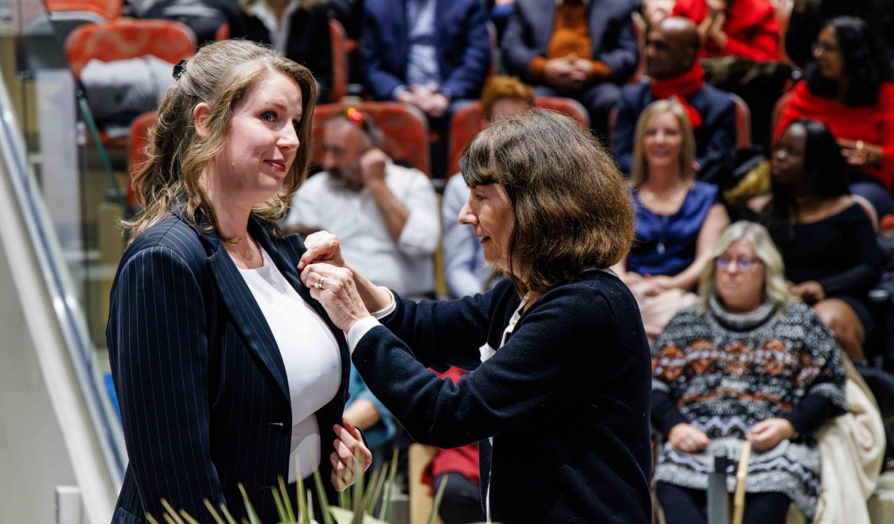 A nursing student, Renata Phillips, wearing a pinstripe blazer, stands on stage as Professor Dawn Prentice attaches a nursing pin to her lapel during Brock University’s Pinning Ceremony on Dec. 13. In the background, attendees watch from tiered seating in the Sean O'Sullivan Theatre.