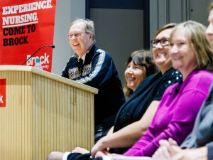 A speaker, dressed in a patterned sweater, addresses a smiling audience during Brock University's Nursing Pinning Ceremony. A red banner in the background reads 