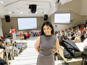 A woman in a grey dress stands smiling at the top of a stairway in a large, wide auditorium at Brock University on Friday, Dec. 13.