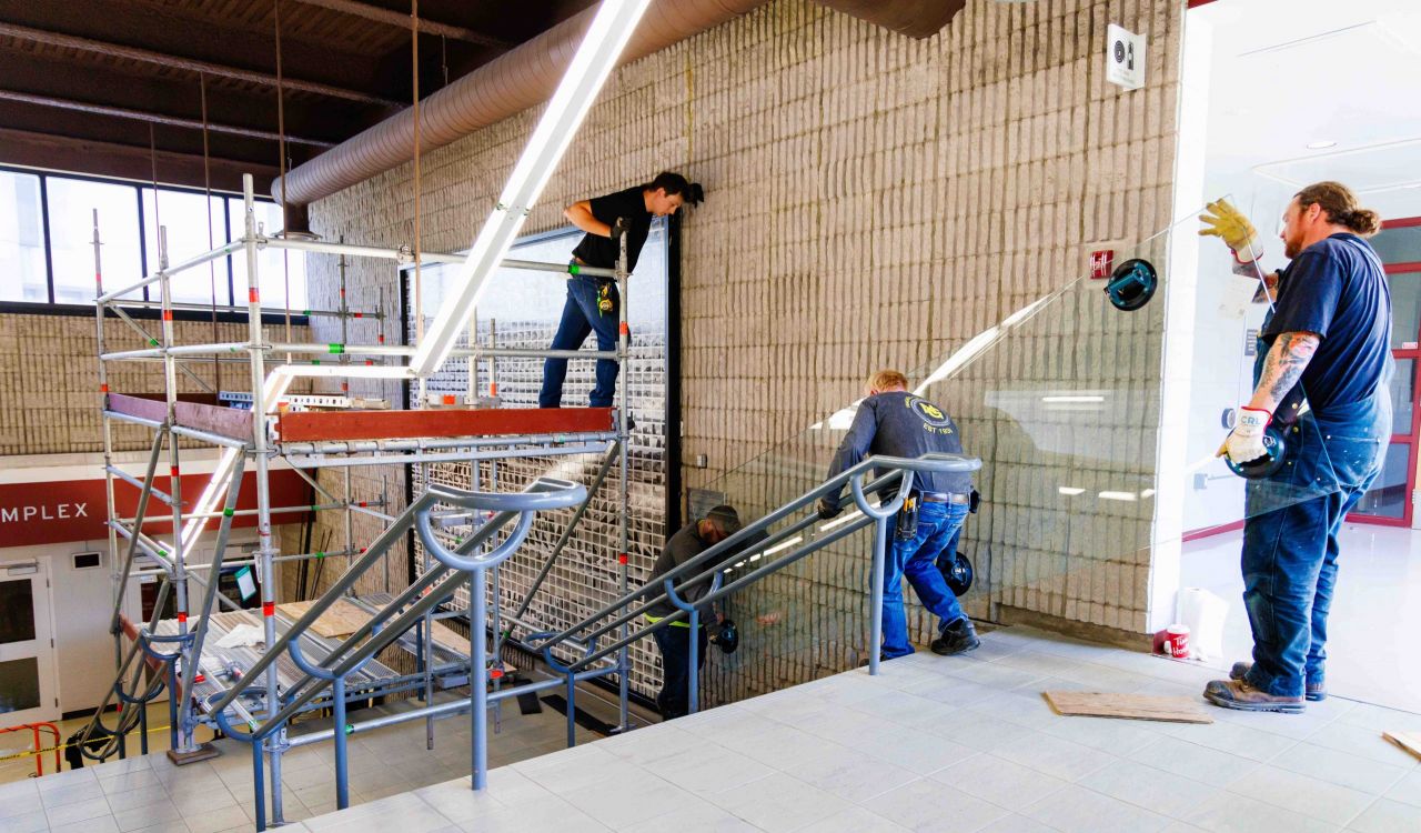 Three men bring in large sheets of glass doing maintenance work on a large mural of photographs hanging in a stairwell.