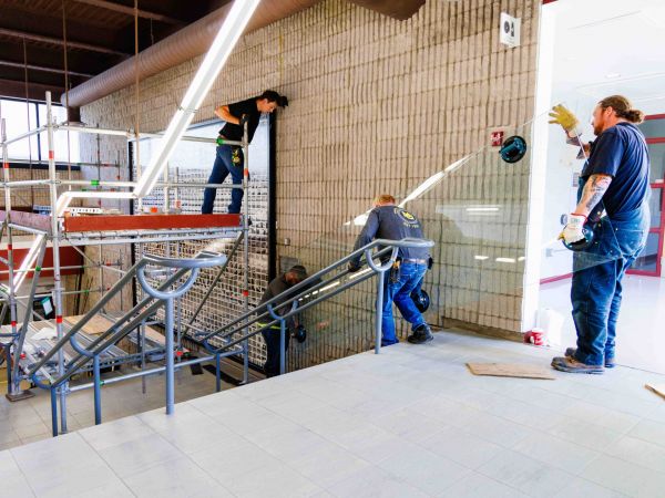 Three men bring in large sheets of glass doing maintenance work on a large mural of photographs hanging in a stairwell.