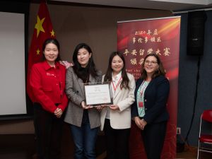 Four women in business casual attire pose for a photo with two of the women holding a glass-framed certificate. A Chinese flag and banner in the background.