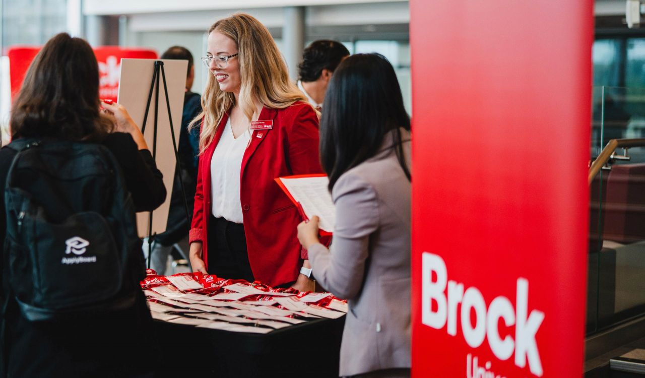 A smiling Brock University representative engages with attendees at a campus event, offering a warm welcome and helpful information.