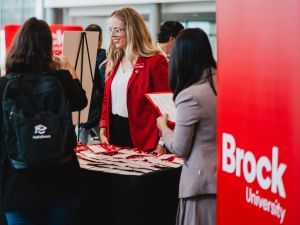 A smiling Brock University representative engages with attendees at a campus event, offering a warm welcome and helpful information.