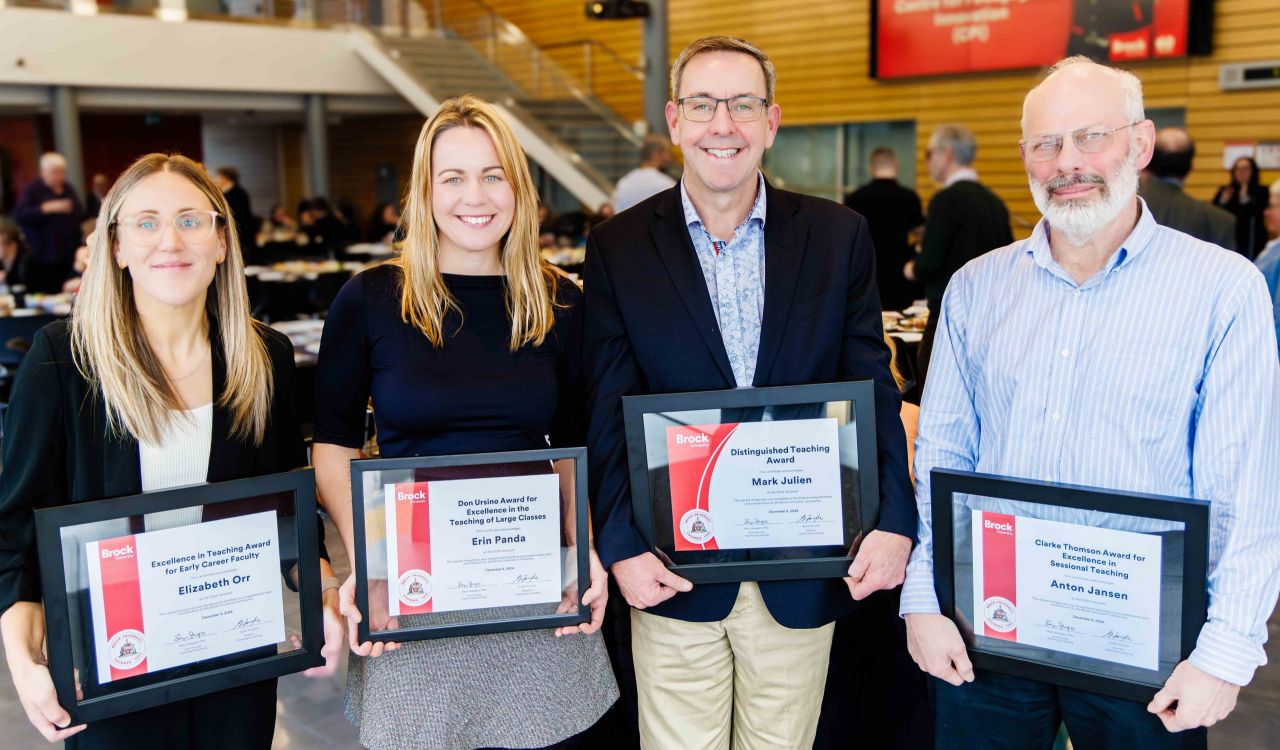 Four award recipients from Brock University stand proudly holding their framed certificates in recognition of their teaching excellence. From left to right: Elizabeth Orr, Erin Panda, Mark Julien and Anton Jansen, each smiling warmly in a brightly lit event space filled with attendees in the background.