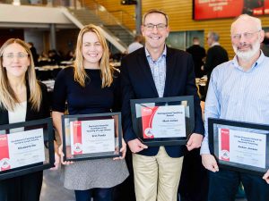 Four award recipients from Brock University stand proudly holding their framed certificates in recognition of their teaching excellence. From left to right: Elizabeth Orr, Erin Panda, Mark Julien and Anton Jansen, each smiling warmly in a brightly lit event space filled with attendees in the background.