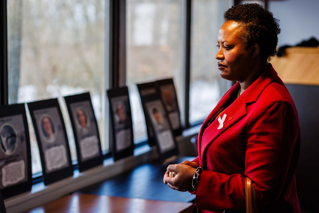 Nadine Williams, dressed in a vibrant red blazer adorned with a white ribbon symbolizing the fight against gender-based violence, stands in quiet reflection at Brock University’s memorial for the National Day of Remembrance and Action on Violence Against Women. In the background, framed tributes to the 14 women killed in the 1989 École Polytechnique massacre line the windowsill, honoring their memory and calling for continued action against gender-based violence.