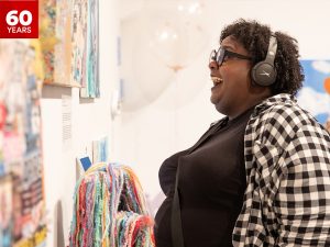 A woman stands in a bright and colourful art gallery looking at a painting, smiling and wearing headphones.