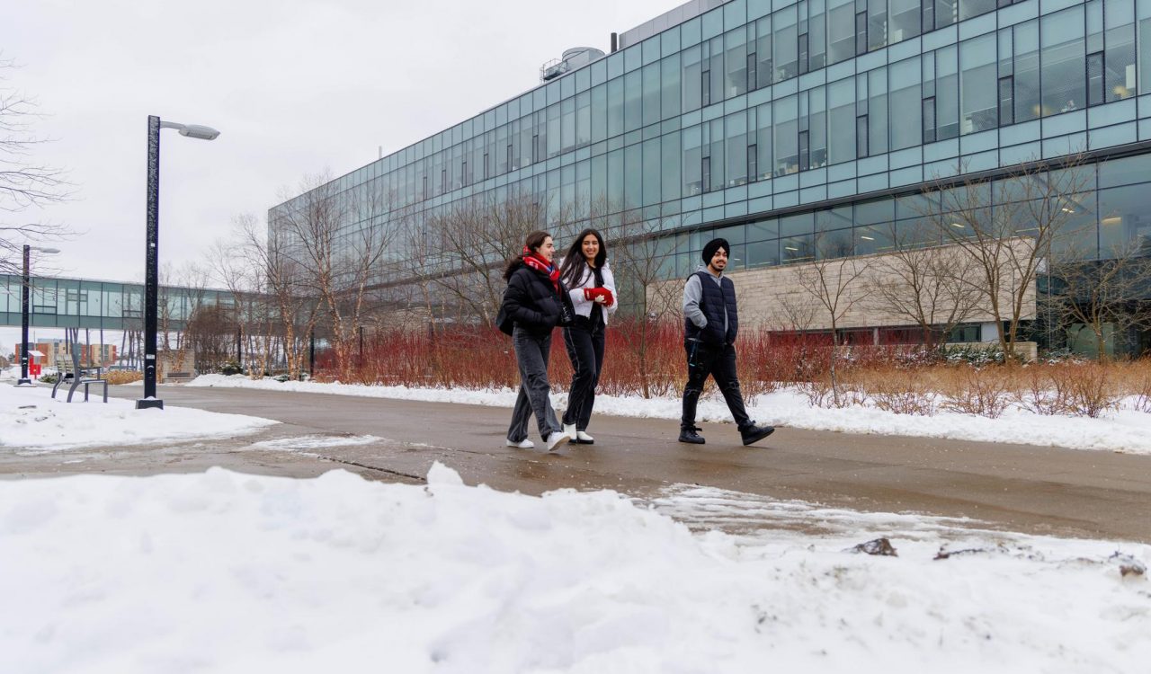 Three people walk on a cleared sidewalk on a snowy day.